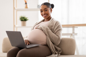 Charming pregnant black woman relaxing with her laptop on bed