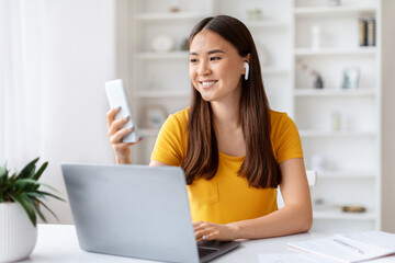 Smiling Asian woman multitasking with smartphone and laptop in home office