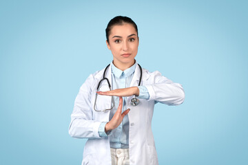 Confident woman doctor in white coat with stethoscope doing time out gesture with hands, blue...