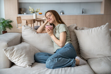 Naklejka na ściany i meble Happy merry middle-aged European woman sitting on comfortable sofa with eyes closed, dreaming, thinking of pleasant things, drinking hot tea, coffee, cocoa, smelling aroma, relaxing in cozy atmosphere