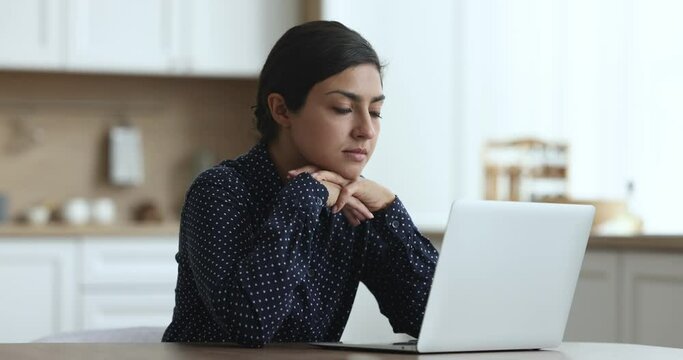 Thoughtful positive young Indian freelancer woman using laptop at kitchen table, working at home workplace, thinking on Internet project at computer, typing, making decision, looking at display
