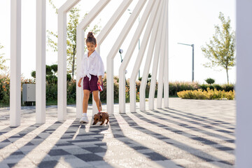 beautiful little girl with long hair walks with a dachshund dog in the summer in the city park - obrazy, fototapety, plakaty