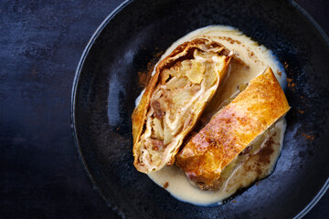 Traditioneller bayerischer Apfelstrudel mit gebackenen Äpfeln, Rosinen und Vanillesoße serviert als Draufsicht auf einem Design Schale