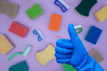 Thumbs up gesture on defocused background of cleaning items. Hand in  blue rubber glove on backdrop of kitchen sponges and dish brushes. Kitchen organization and cleaning.