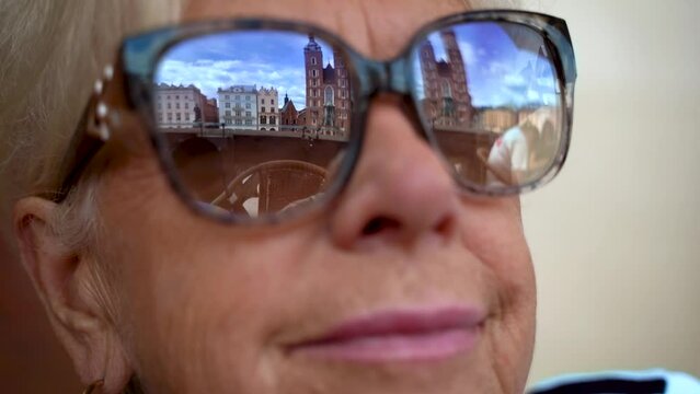 The historic market square in Krakow with St. Mary's Basilica reflected in sunglasses. A figure reflected in glasses waving at the viewer. Face of a mature woman in sunglasses.