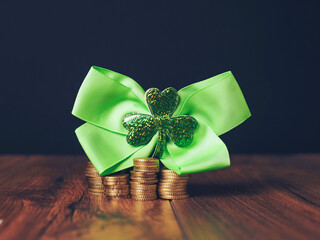 Green ribbon with shamrock symbol of Ireland on pile of coins on wooden brown surface, dark...