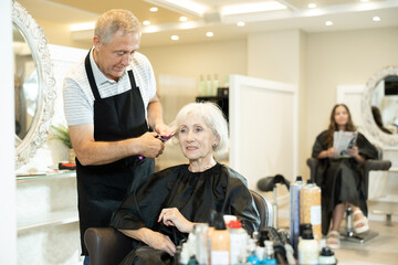 Elderly male hairdresser performs hair styling and creates hairstyle with curling iron and comb. Elderly female client is resting during beauty procedure