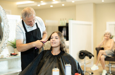 Elderly man, professional experienced hairdresser using curling irons to create curls and add volume to hair of young interested smiling girl..