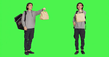 Standing over full body greenscreen, asian guy operates on food delivery with heated backpack. Young man holding bag for takeaway, prepared to give meal orders to customers.