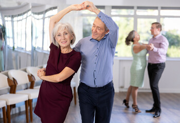 Senior man and elderly woman are dancing classic version of waltz in couple during lesson at studio. Leisure activities and physical activity for positive people.