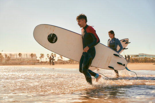 Side view of surfers in wetsuits running into the sea holding surfboards - Powered by Adobe