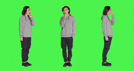 Adult doing hush secret sign over greenscreen, expressing secrecy symbol with finger over lips. Young person showing mute silence gesture , being private in studio with full body backdrop.