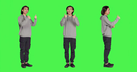 Obraz premium Asian guy does thumbs up gesture standing over full body greenscreen, showing agreement and presenting like symbol in studio. Young joyful adult feeling pleased, approval sign.