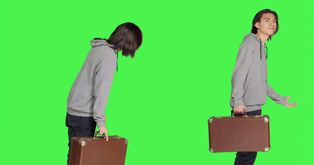 Young adult holding vintage suitcase over greenscreen backdrop, waiting impatiently for a certain thing. Asian man carrying huge briefcase while he looks at time on wrist watch in studio.