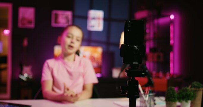 Charming small child shooting video for social media platform in living room, talking about her day. Kid entertaining followers, doing content creation in pink neon lit apartment using phone on tripod