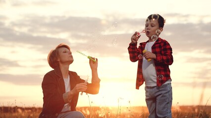 Mother and son blow soap bubbles together at back sunset in middle of field in countryside overlooking beauty of nature. Mother entertains son walking in nature. Fun from mother for son
