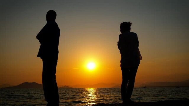 Sad couple silhouette back to back on beach. A view of stressed couple silhouette standing on the beach against orange evening sky after quarrel.