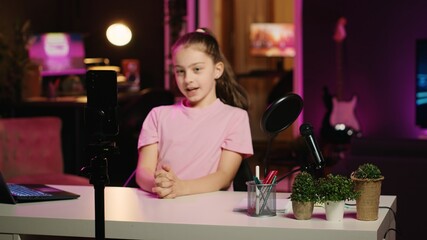 Young girl filming content for her online fans, talking about gen Z topics. Cute child recording vlog for family channel in apartment studio with pink neon lighting as background