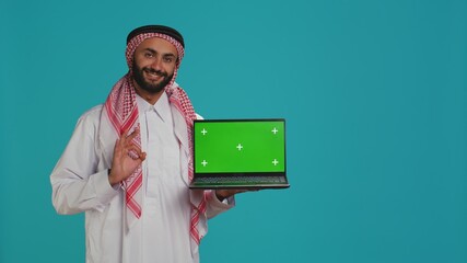 Muslim adult shows greenscreen laptop with blank mockup layout on display, copyspace template. Arab guy wearing gown and headscarf, presenting isolated chromakey screen on pc.