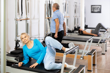 Focused man and two women of different ages doing stretching exercises while using Pilates bed machine in rehabilitation center