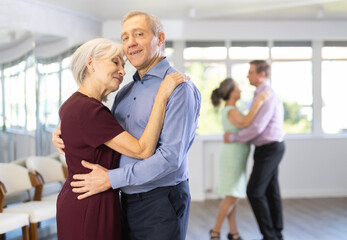 Smiling man supporting mature lady, dancing slow foxtrot in pair during group class in modern...