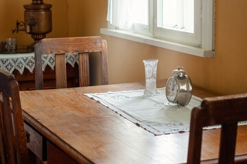 An antique wooden table and three chairs by the window, daylight streaming in to illuminate a baptismal vase and a large antique clock on the table.
