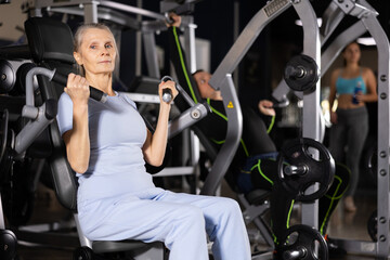 Fototapeta na wymiar Focused elderly woman doing strength training in gym, exercising on shoulders press machine..