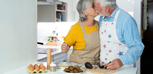 Happy joyful older husband and wife kissing togetherness, making organic fresh salad together, talking, laughing. Old couple having fun in kitchen, preparing dinner.