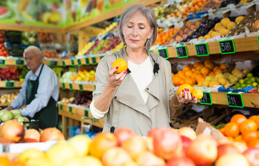 Obraz premium Old lady choosing fruits while standing in salesroom of greengrocer. Senior merhcandiser working in background.