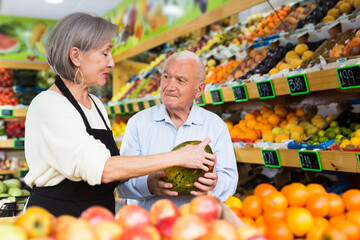 Mature woman greengrocer worker helping old man to choose fresh melon.