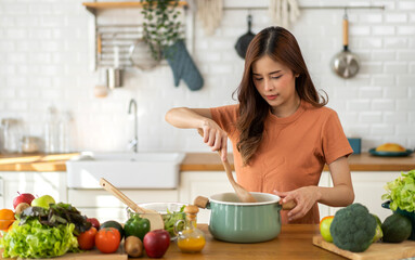 Young woman standing near stove and cooking, housewife, meal, chef, food.Happy woman looking and smelling tasting fresh delicious from soup in a pot with steam at white interior kitchen.