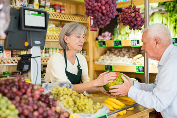 Senior saleswoman standing at counter in greengrocer and serving man customer who purchasing fresh...