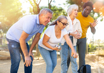A group of multiracial middle aged mixed-sex adult people playing petanque game outdoors in public...