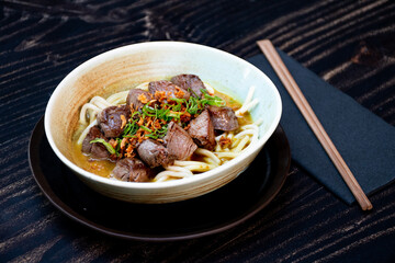 dish of oriental noodles with cubes of grilled meat on a wooden table with chopsticks in close-up