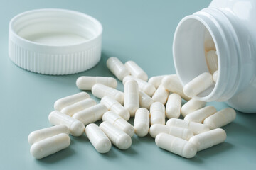 White medical capsules next to open plastic container - bottle on pastel green-blue background. Photo. Selective focusing