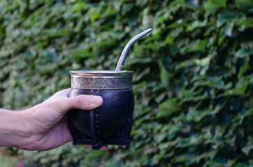 Young man's hand holding a yerba mate to share on green nature background.