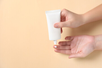 Woman applying cosmetic cream from tube onto her hand on beige background, top view. Space for text