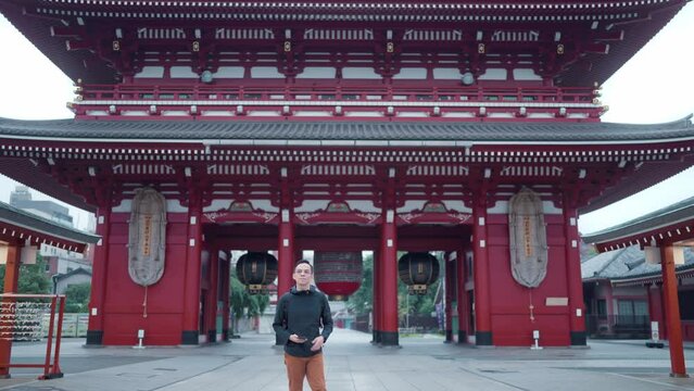 View full body of hispanic tourist guy standing on blurred background of Japanese temple Sensoji and taking photos with mobile phone during journey in Asakusa Tokyo, Japan