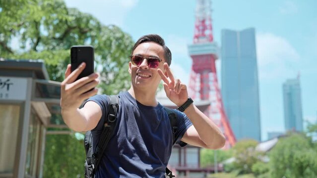 Smiling young Hispanic male traveler in casual wear and sunglasses smiling and taking selfie with smartphone while standing against ancient Zojo ji Buddhist temple near Tokyo tower, Japan