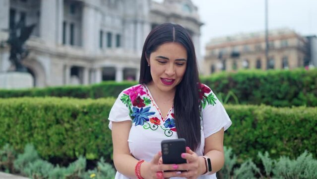 Happy young ethnic mexican woman using smartphone and looking at screen of mobile phone while sitting in front of blurred Bellas Fine Arts Palace in Mexico City and wearing traditional clothes