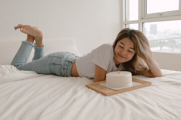 Lovely beautiful happy asian woman lies on the bed admiring her birthday cake.
