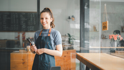 Beautiful young barista woman in apron holding tablet and standing in front of the door of cafe...