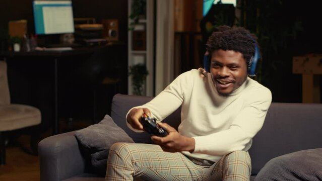 Gamer wearing headphones, feeling happy after receiving winner screen on gaming console for killing all enemies. African american player at home bragging after being victorious in videogame