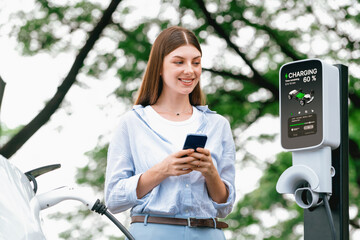 Young woman using smartphone online banking application to pay for electric car battery charging...