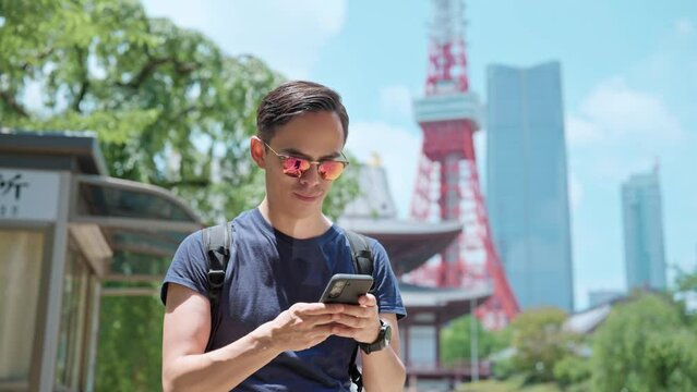 Smiling young Hispanic male traveler in casual wear and sunglasses with backpack smiling and messaging on smartphone while standing against ancient Zojo ji Buddhist temple near Tokyo tower, Japan