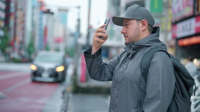Side view of concentrated young Hispanic male tourist in warm clothes and cap with backpack taking photo on buildings on smartphone while standing in Akihabara neighborhood in Tokyo, Japan