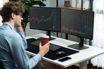 Working young business trader focusing on market stock graph data in two screens, holding coffee...