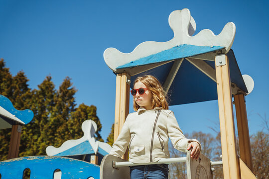 Cute preschooler girl climbing on a playground equipment on the blue sky background. happy girl in beige jacket and sunglasses is playing on playground, outdoor