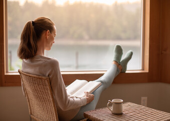 Happy woman in cozy sweater enjoying reading a book and cup of hot drink , sitting next to window...