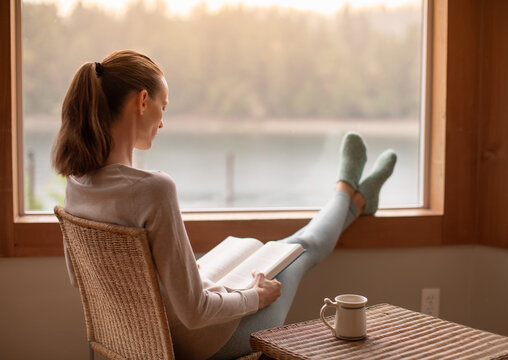 Happy woman in cozy sweater enjoying reading a book and cup of hot drink , sitting next to window enjoying the winter morning at home
